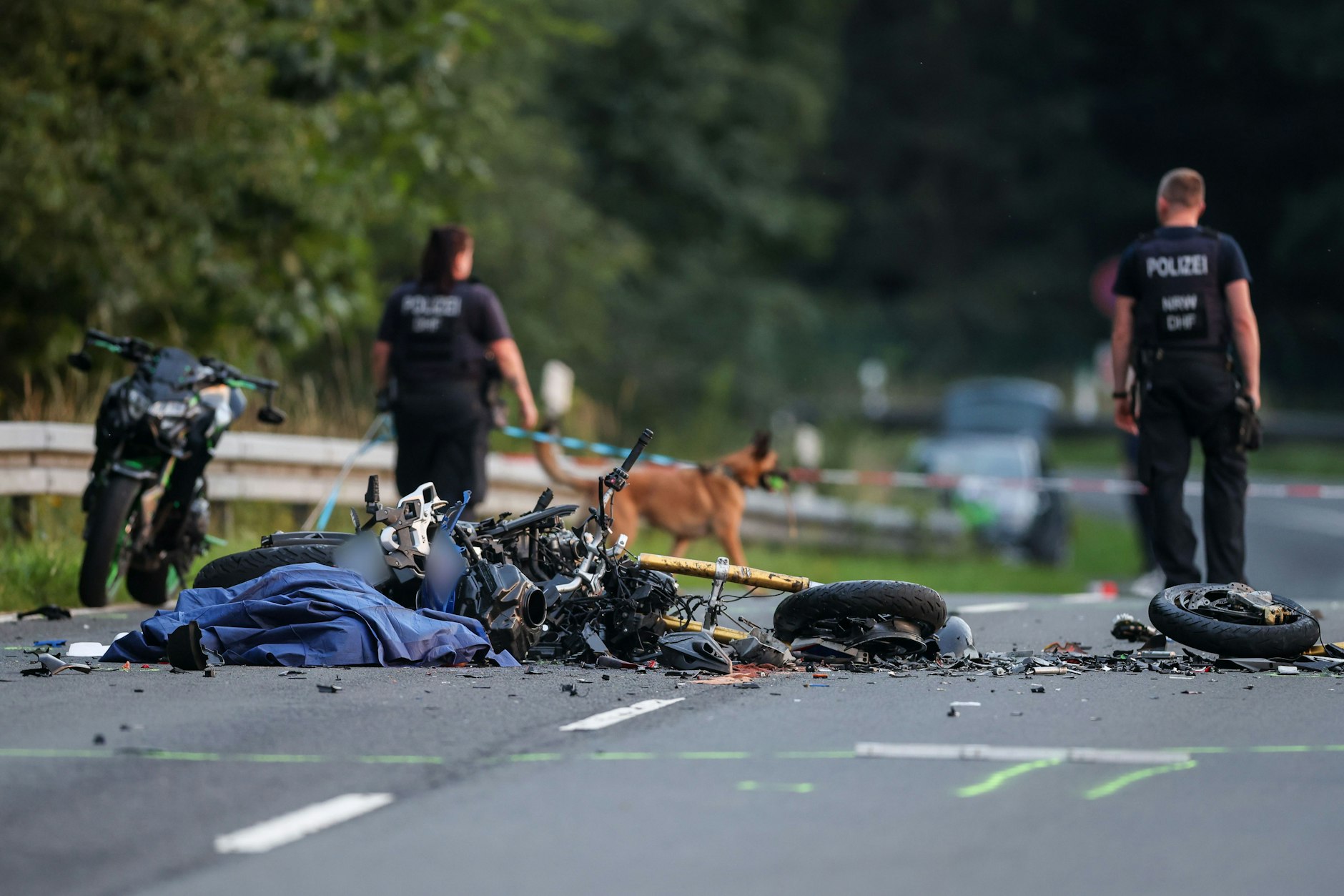 Ein vollkommen zerstörtes Motorrad liegt nach dem Frontalunfall auf der Straße, Polizeibeamte sichern Spuren.