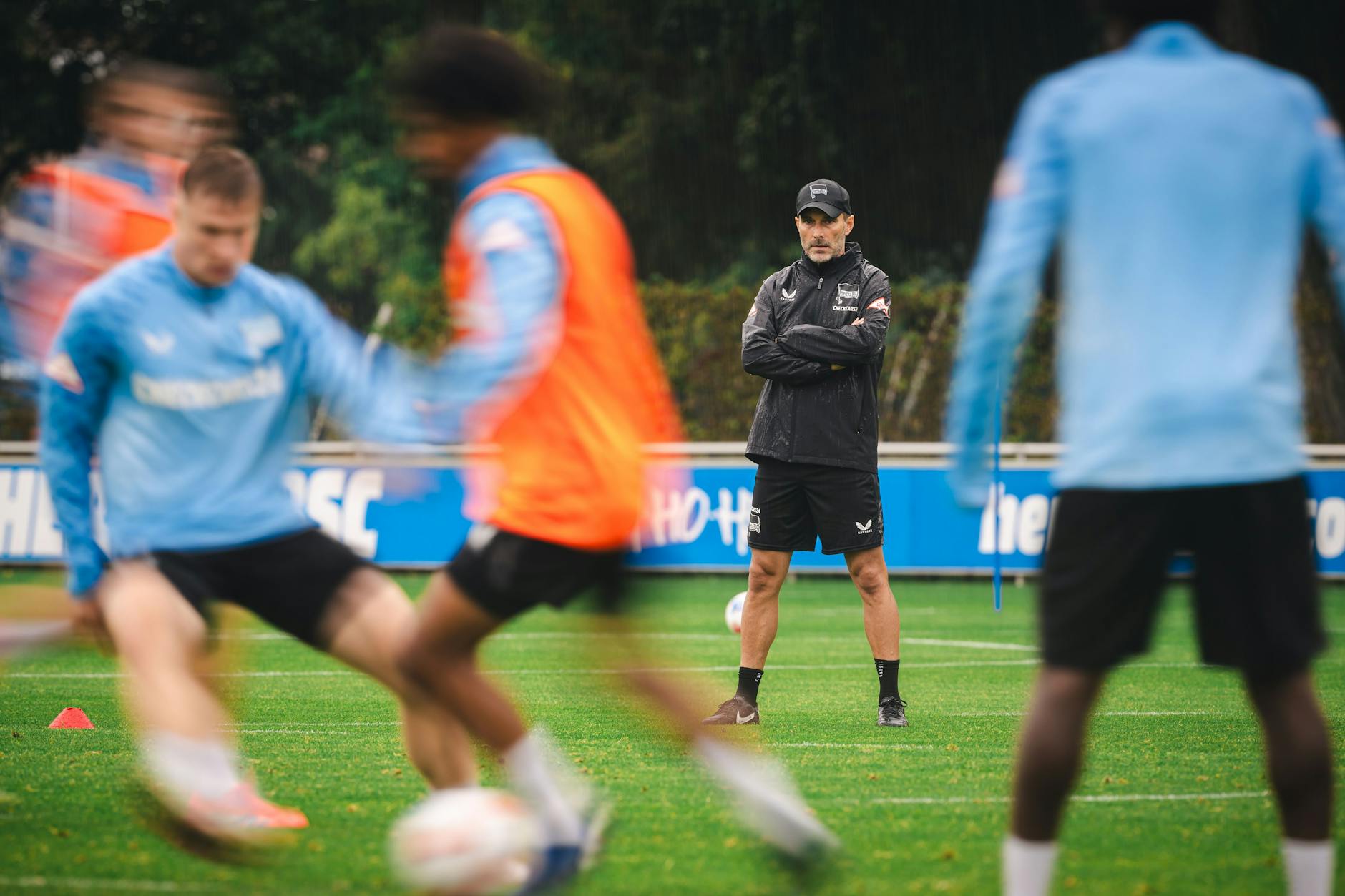 Coach Stefan Leitl steht beim Training von Hertha BSC mit vor der Brust verschränkten Armen auf dem Rasen, schaut den Profis genau auf die Füße.